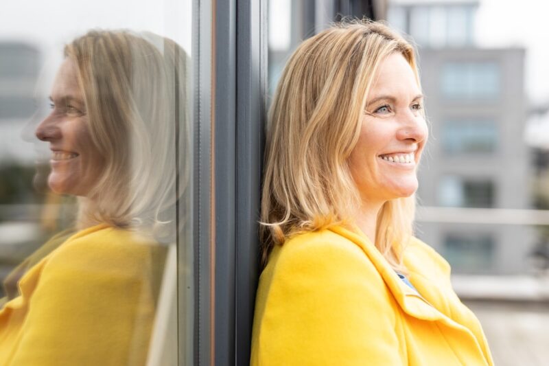 Verena Machnik, in gelber Jacke an Fenster gelehnt (Fotografie: Christian Klant)